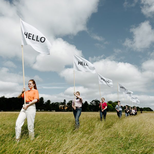 A line of people hold flags reading Hello in the meadow of Jupiter Artland for Edinburgh Art Festival 2020.