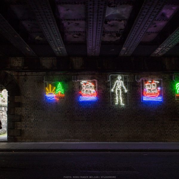 Image of Graham Fagen's work, A Drama in Time, made up of 5 neon illustrations under bridge. The entrance to Jacob's Ladder sits on the left.
