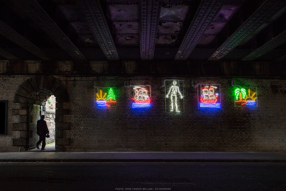 Image of Graham Fagen's work, A Drama in Time, made up of 5 neon illustrations under bridge. The entrance to Jacob's Ladder sits on the left.