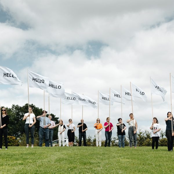 Young people stand in a row holding white flags that read 'HELLO' in field.