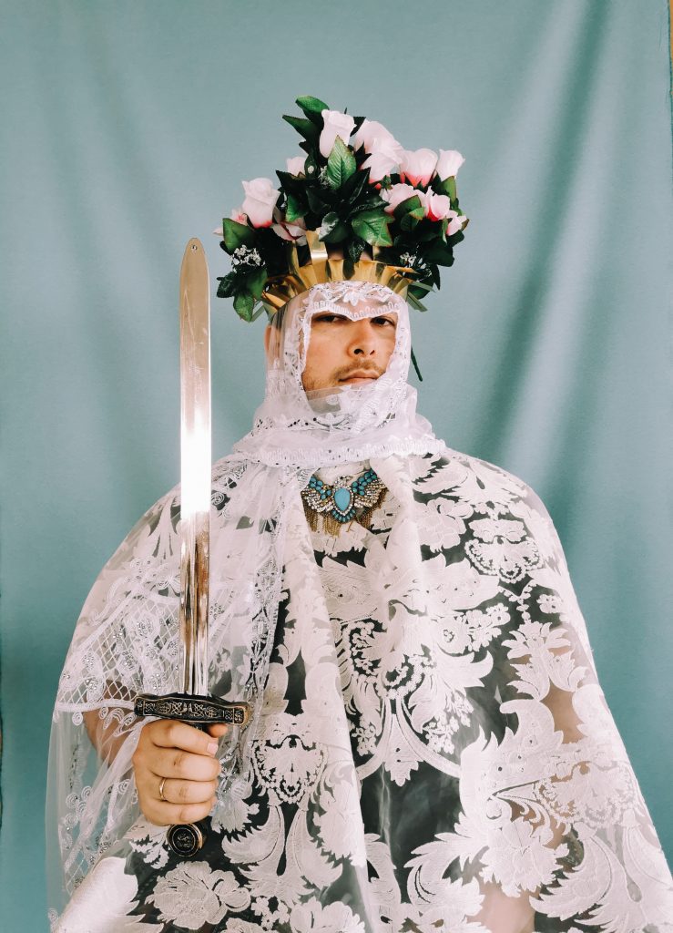 Artist Jose Campos holds sword in floral headdress, against turquoise curtain. 
