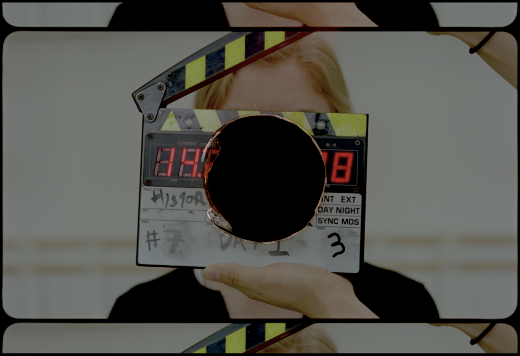 Hand holds clapperboard in film still, with hole burnt through the middle of image of person's face.