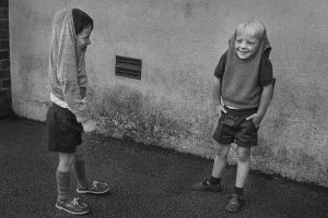 Black and white film photo of two boys with jumpers pulled over their head giggling at each other.