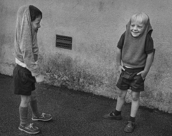Black and white film photo of two boys with jumpers pulled over their head giggling at each other.