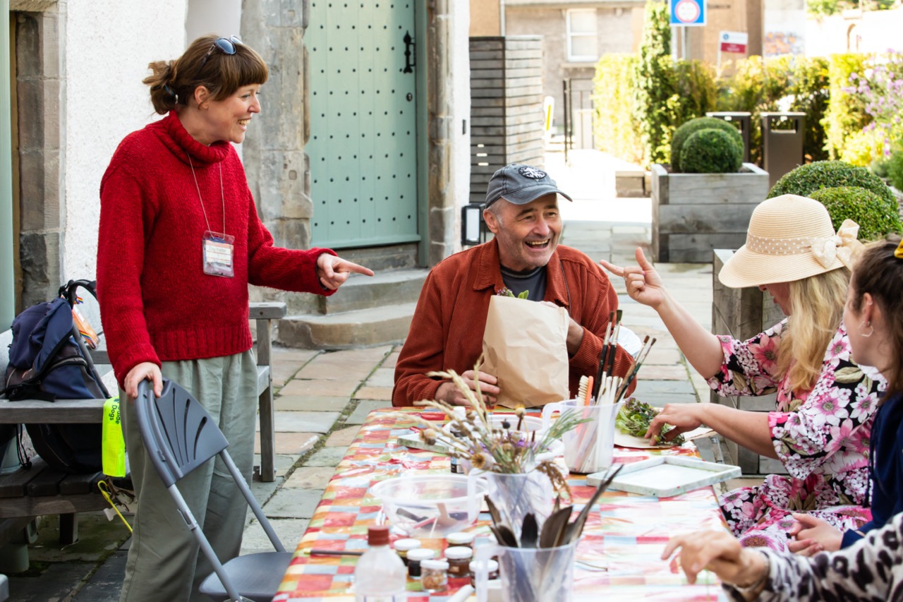 Four people - one standing and three sitting - around an art workshop table in a garden.