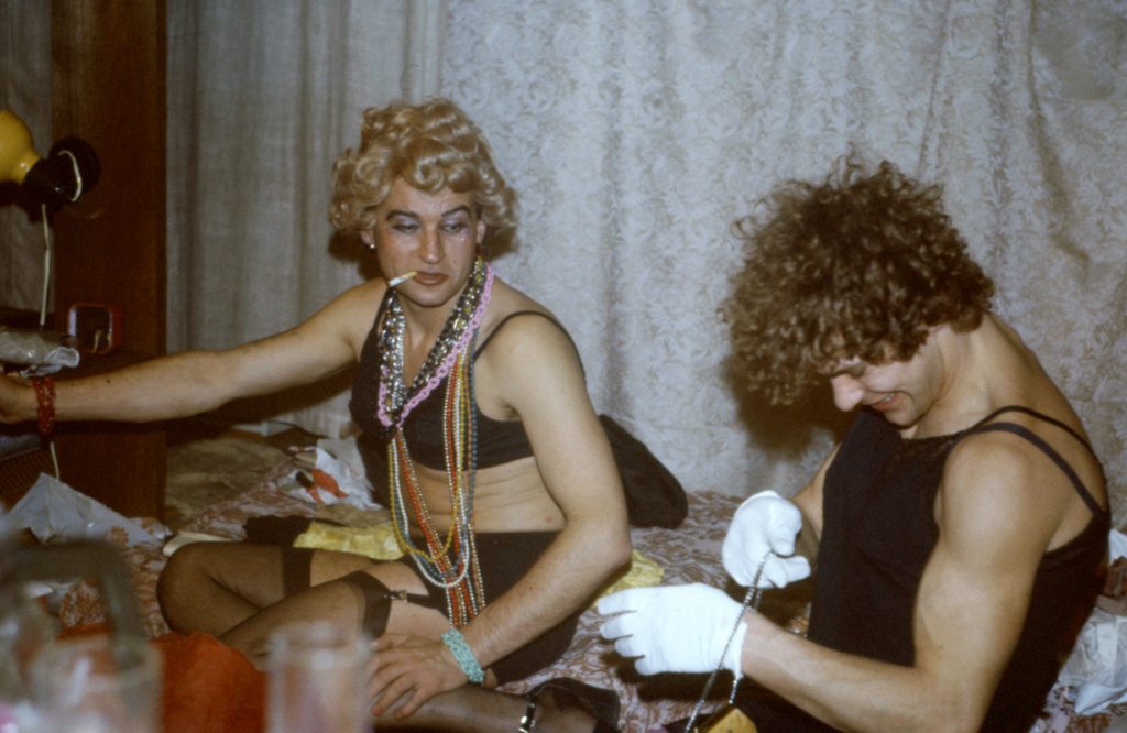 Photo of two men dressed glamorously in lingerie, wigs, stylised makeup and costume jewellery sitting on the floor in front of a hanging curtain.
