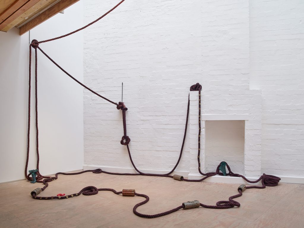 Photo of Unravelled Gathering, an installation by Alaya Ang of a thick rope threaded and draped across a neutral wall and floor, looped with large beads and casts of feet.
 