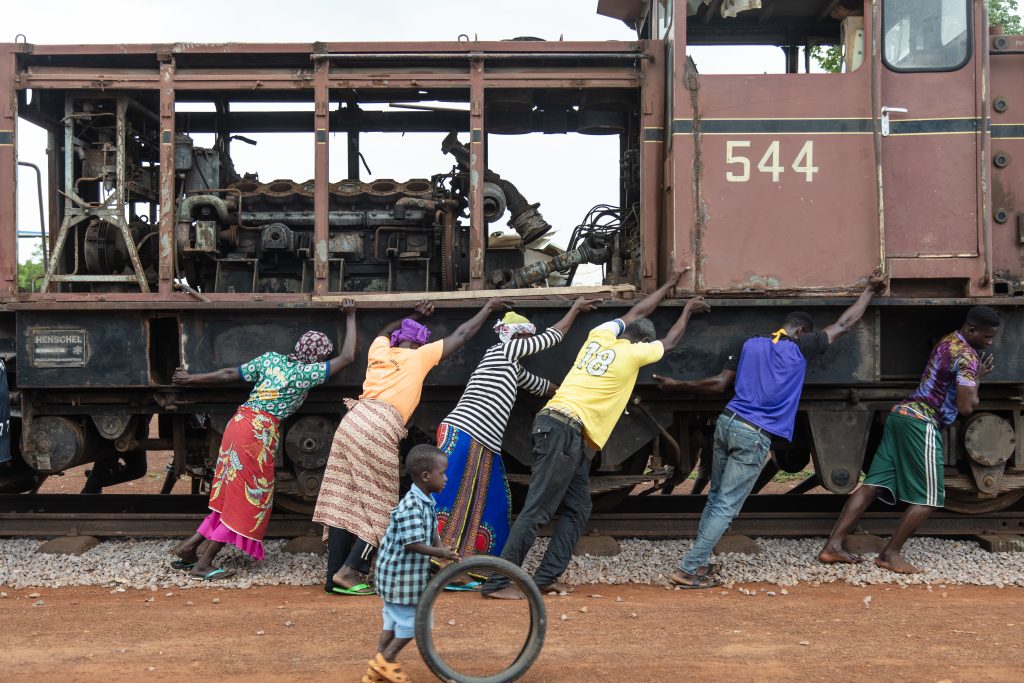 A photo by Ibrahim Mahama, of a row of colourfully dressed people leaning against a stripped train car with visible inner working, as if pushing it along its rails. A child plays with a tyre in the foreground.