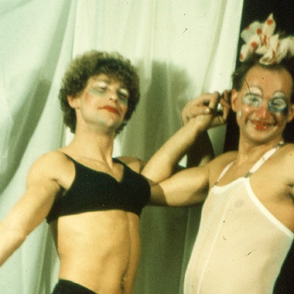 A photo showing two men dressed flamboyantly in a mixture of stylised makeup, lingerie, skirts, and hair bows, posing in front of a hanging sheet.