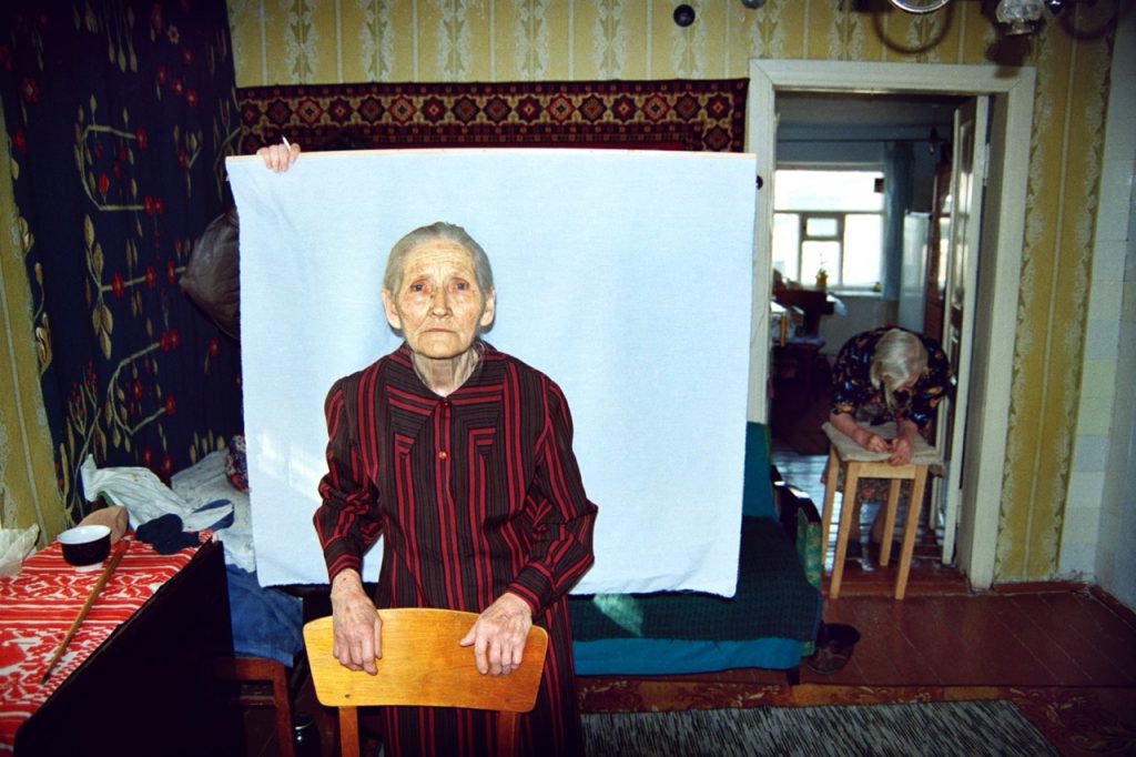 A photo by from Alexander Chekmenev’s Passport series, of an older woman in a domestic setting, looking into the camera in with a white square of fabric held up behind her, as if getting a passport photo taken.
 